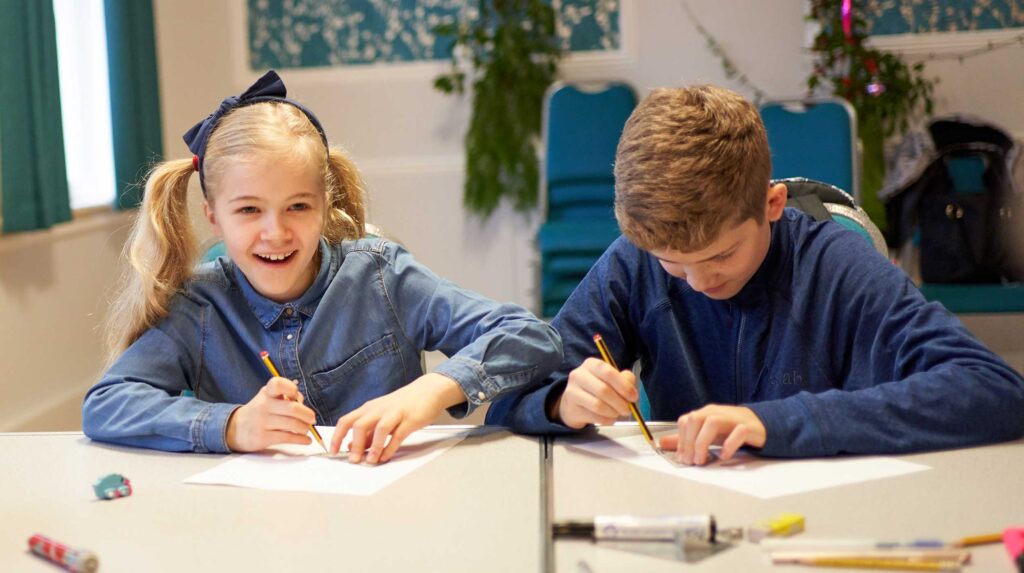 Dwoje uśmiechniętych dzieci rysujących ołówkami w otoczeniu klasy szkolnej